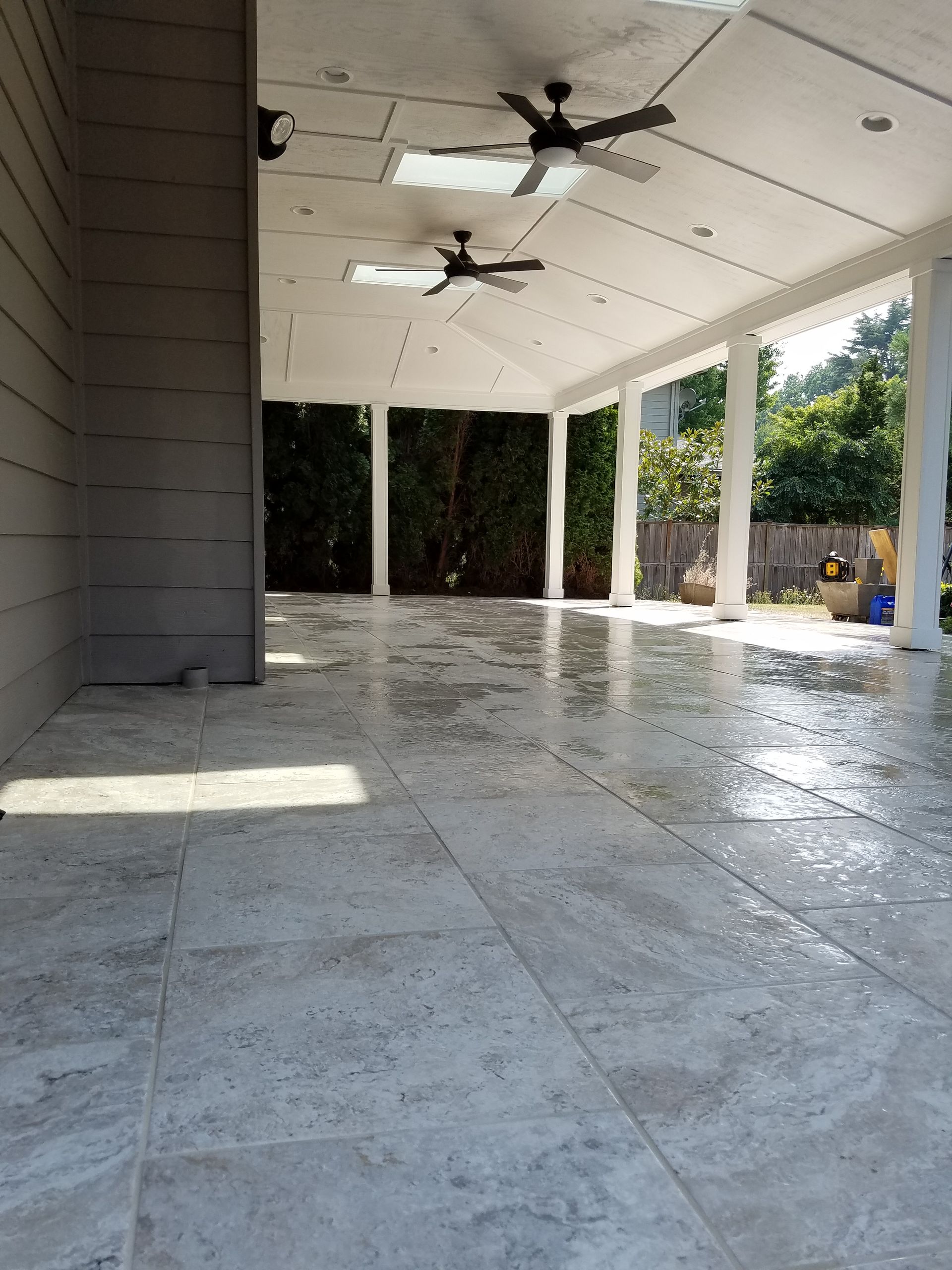 Covered patio with light-colored tile flooring, open to a backyard with trees. Ceiling fans and recessed lights.