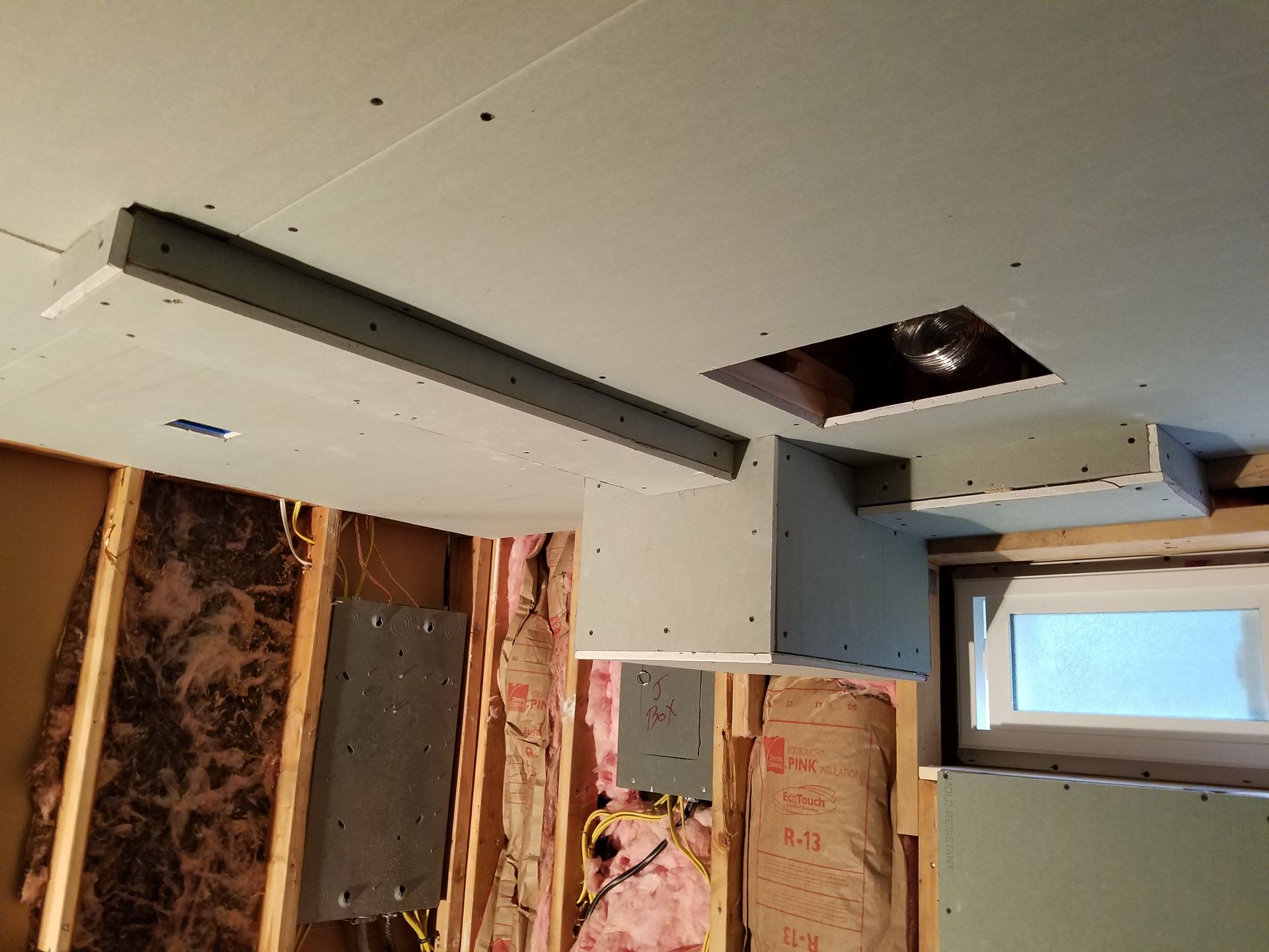 Drywall installation on a ceiling with a built-out structure. The walls are framed with insulation and a window.