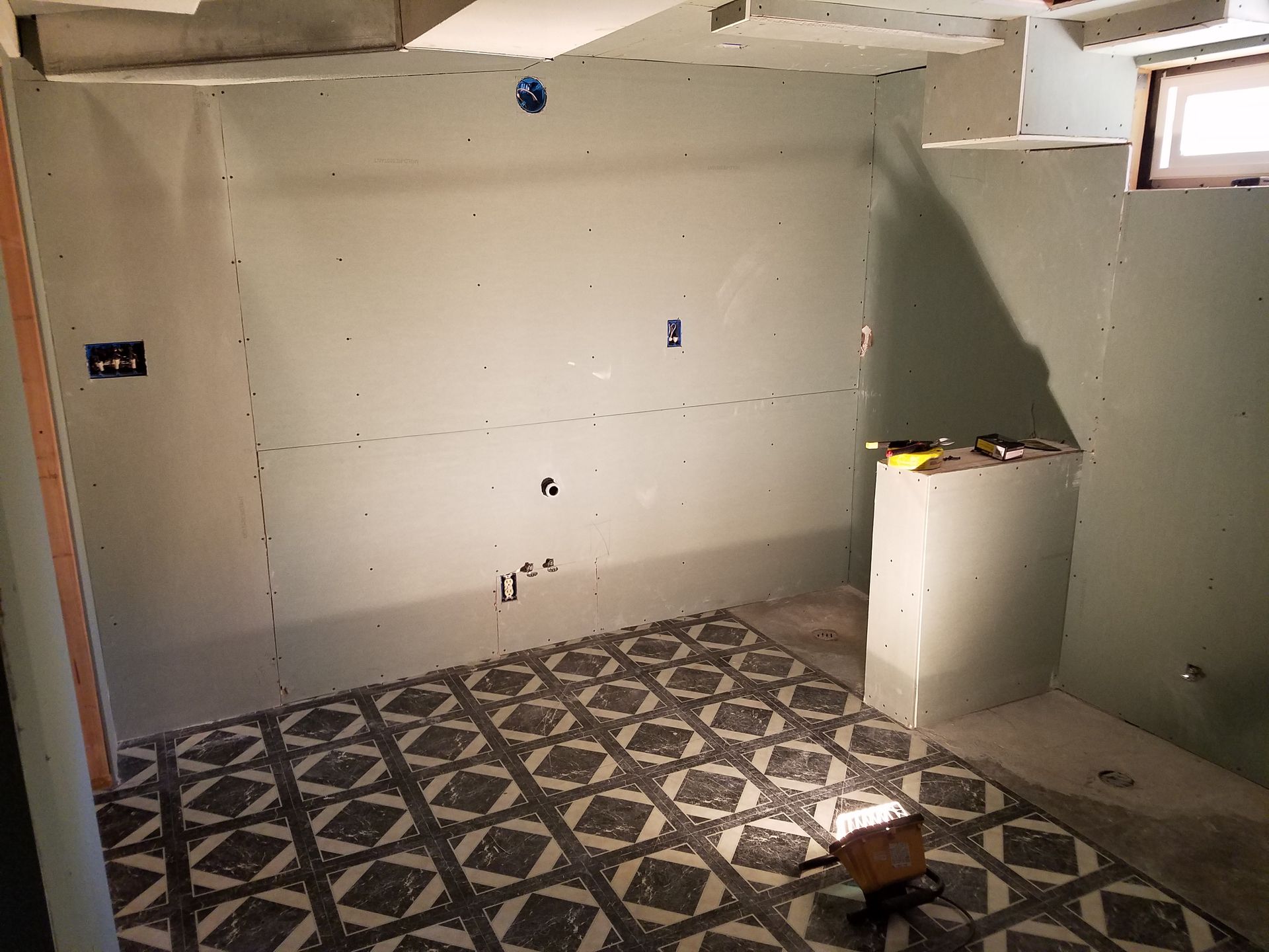Bathroom under construction; walls of greenish drywall, patterned floor, small window.