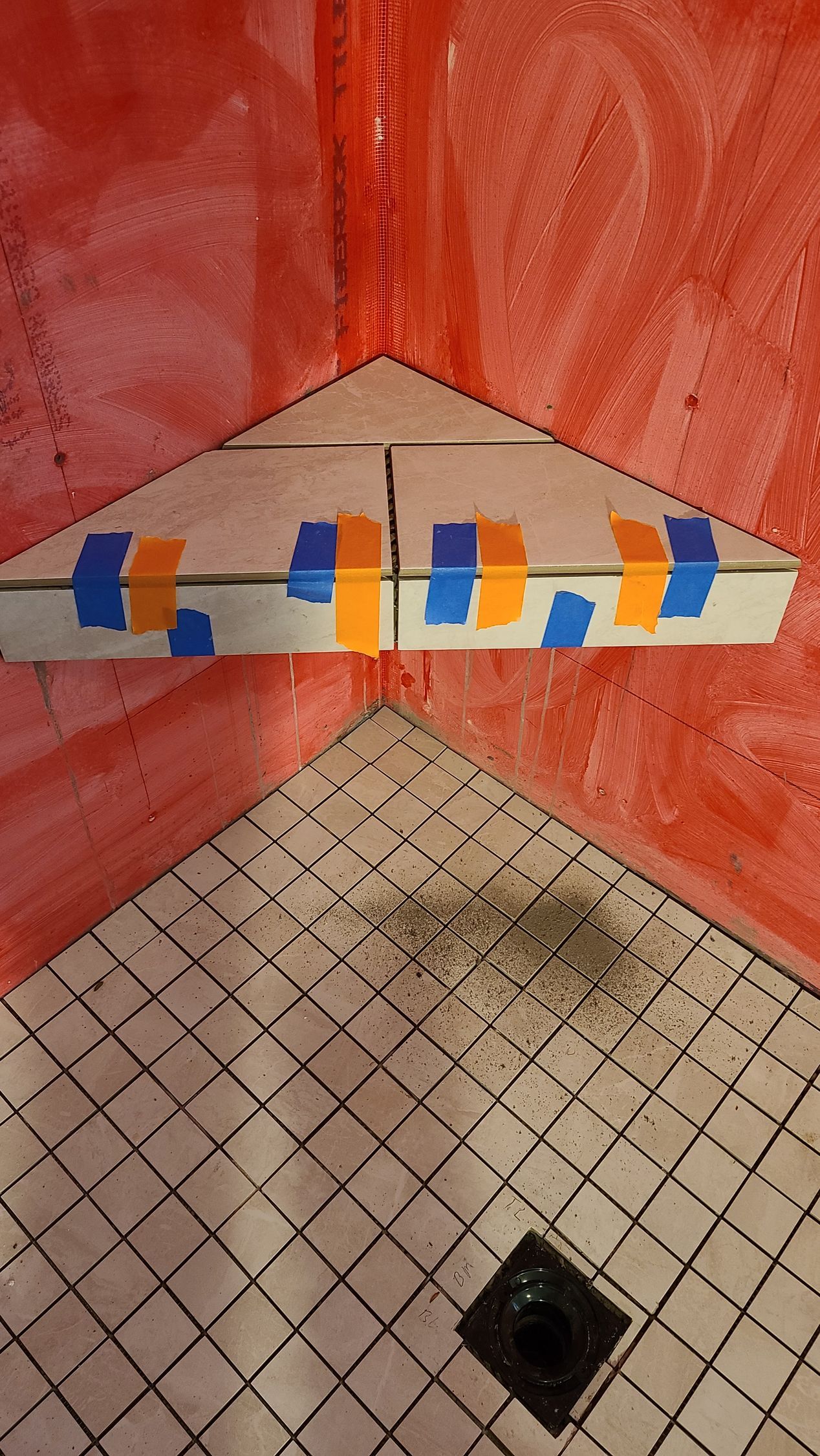 Shower floor with tiles and a triangular shelf taped in place, red waterproof sealant.