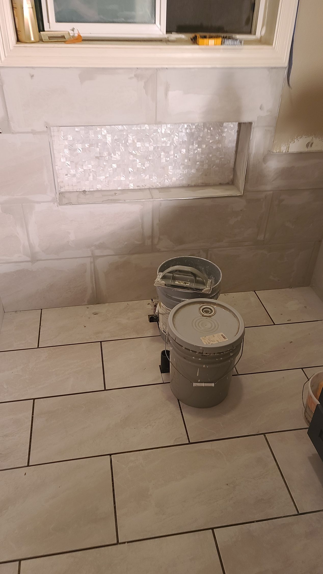 Bathroom under construction, with tiled floor, cinder block walls, and buckets. A window is above.