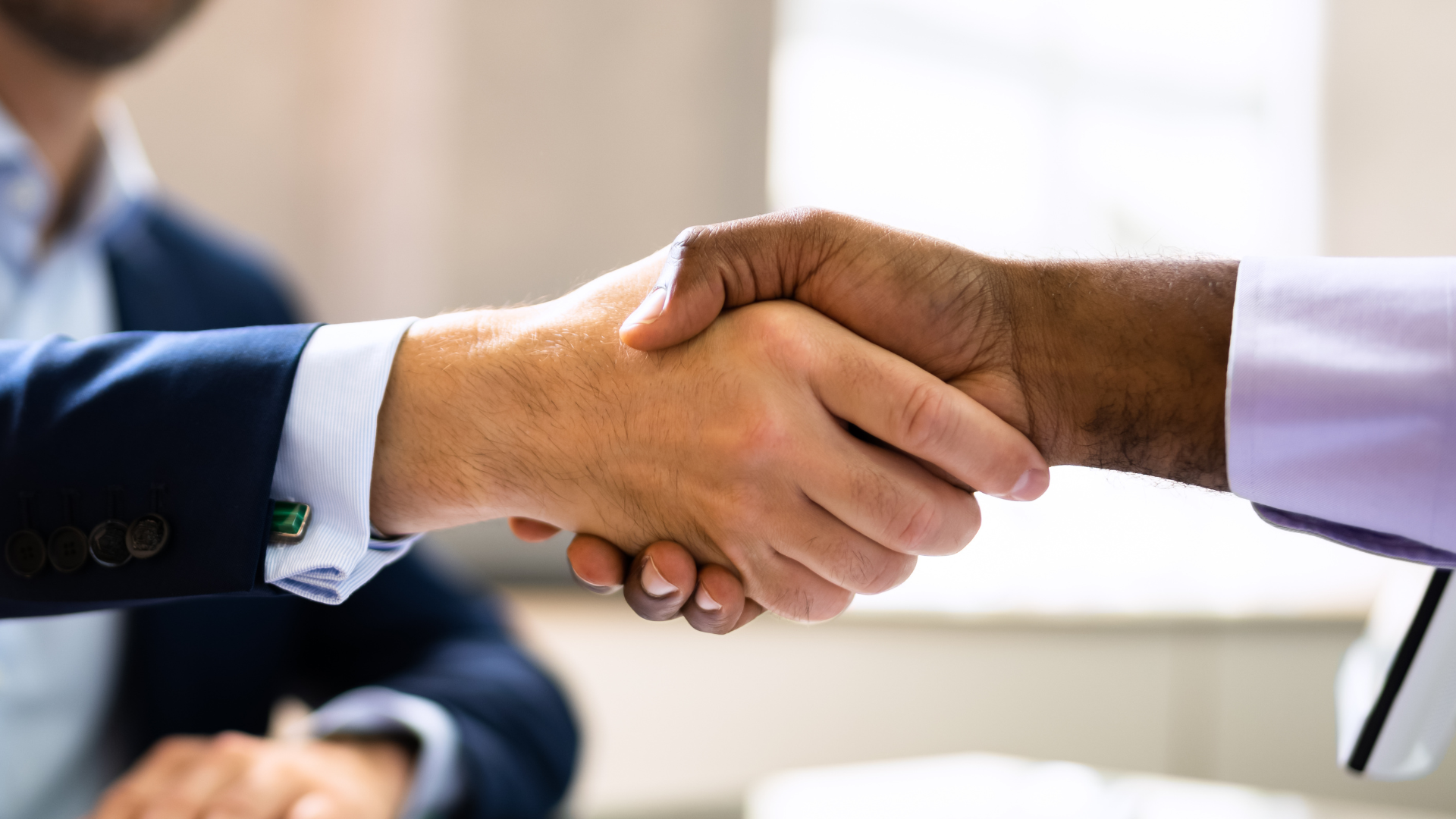 Two people shaking hands, one in a blue blazer, the other with brown skin in a purple shirt, indoors.