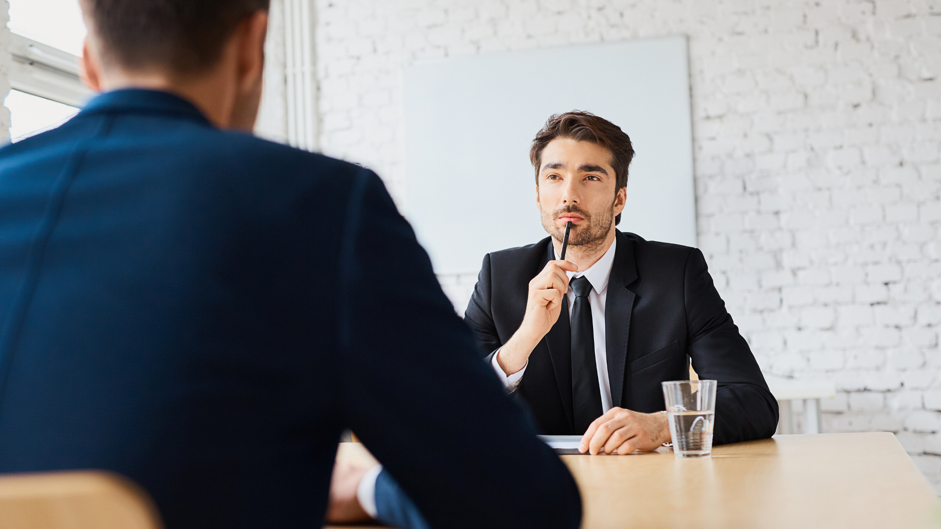 Man in suit interviewing another man, holding pen, looking thoughtful.