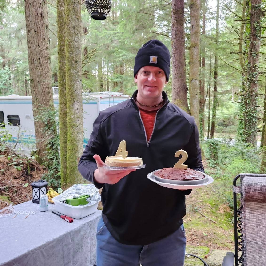 Man outdoors in a forest holding two dessert plates with number candles, standing by a campsite table