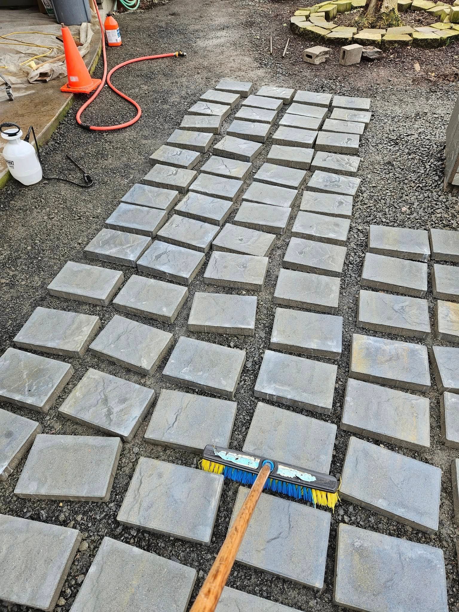 Workers laying gray pavers on a gravel path with orange cones and tools nearby