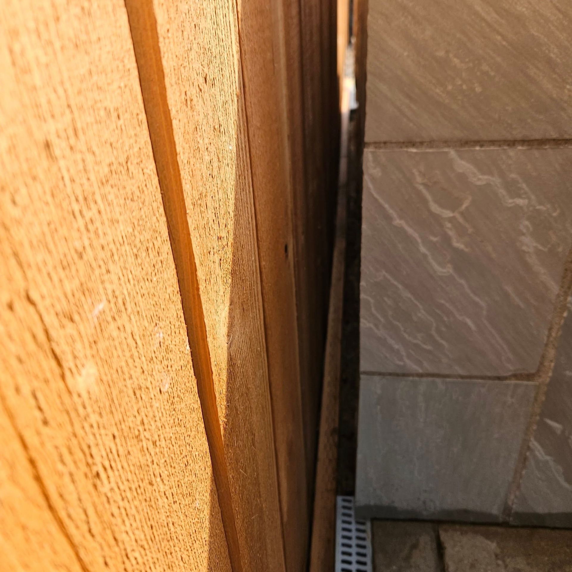 Close-up of a narrow gap between a wooden wall and stone tiles, with a drain grate at the bottom.