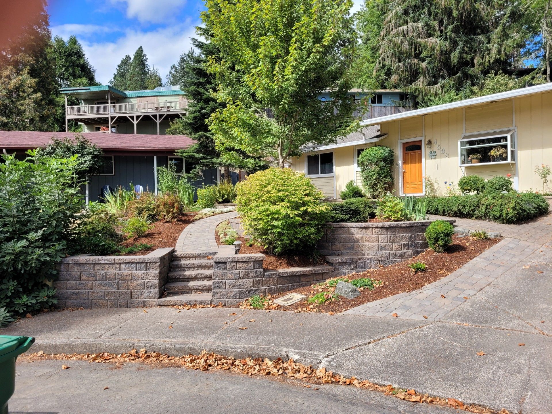 Courtyard garden with curved driveway, stone retaining walls, and two modern buildings under tall trees