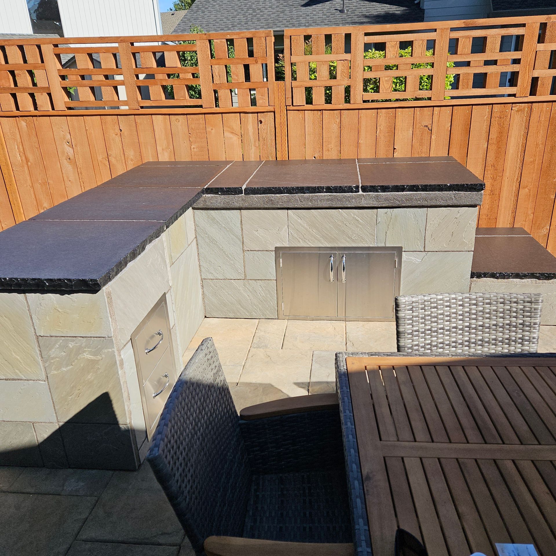 Stone outdoor kitchen and patio with dark countertops, wooden fence, and woven chair in sunlight