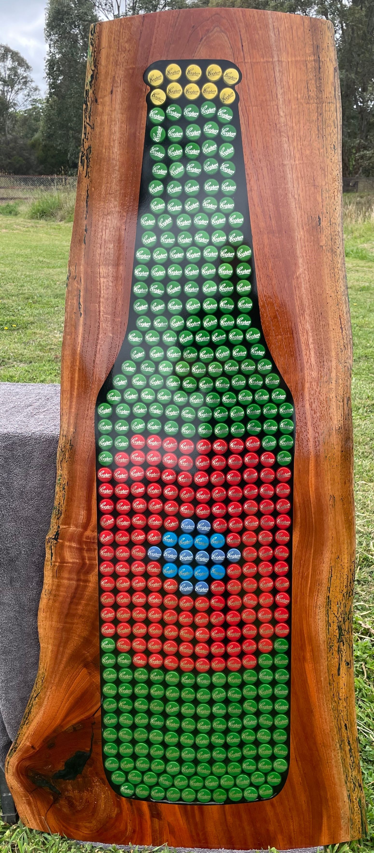 A bottle shape made of bottle caps embedded in wood. It has green, red, yellow and blue caps.