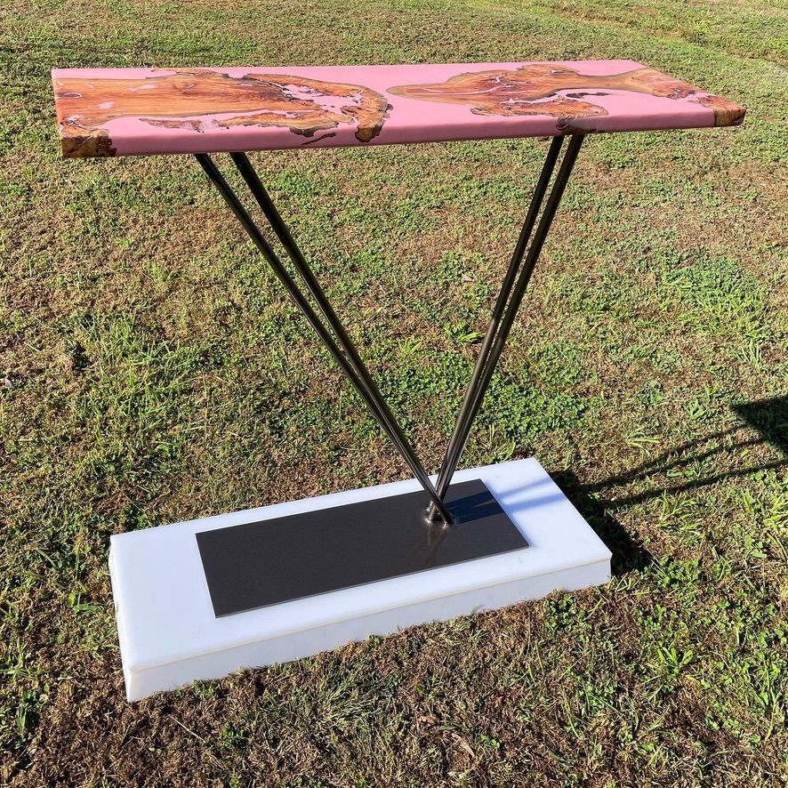 Pink resin and wood table on metal legs, over a white base, on grass.