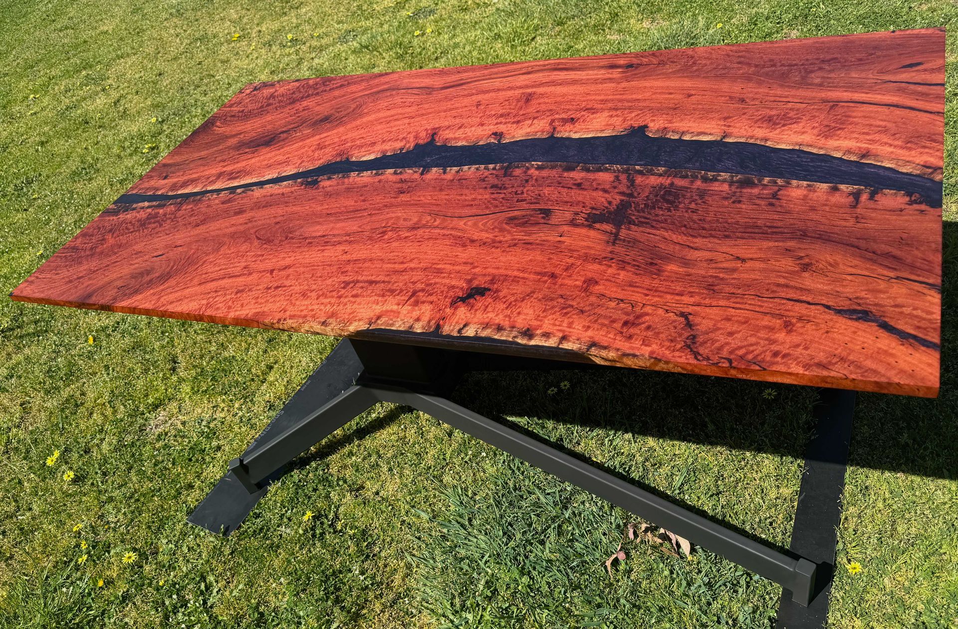 Reddish-brown wood table with black epoxy river, on black metal legs, set on green grass.