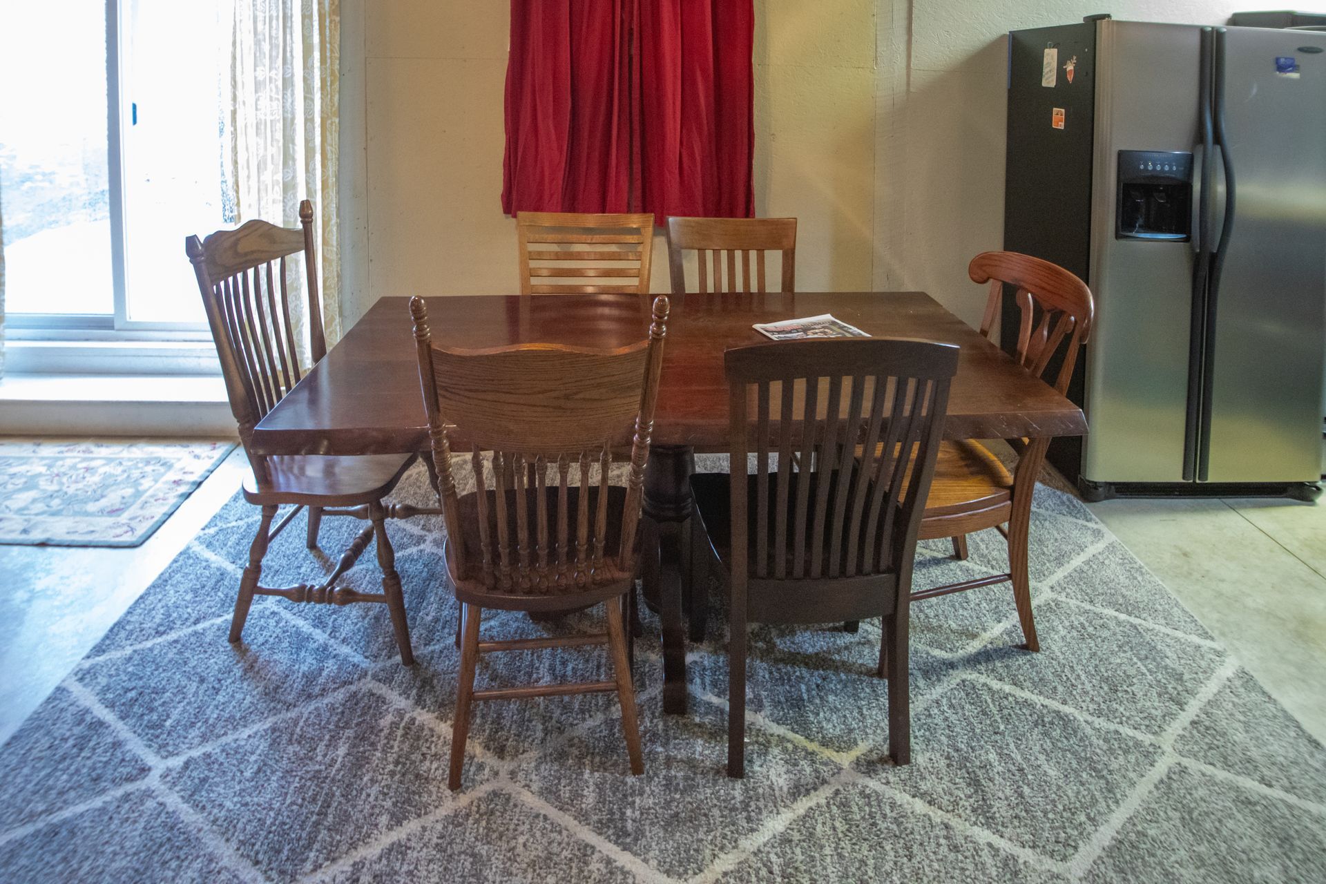 A dining table with six wooden chairs, under a red curtain and next to a refrigerator, on a patterned rug.