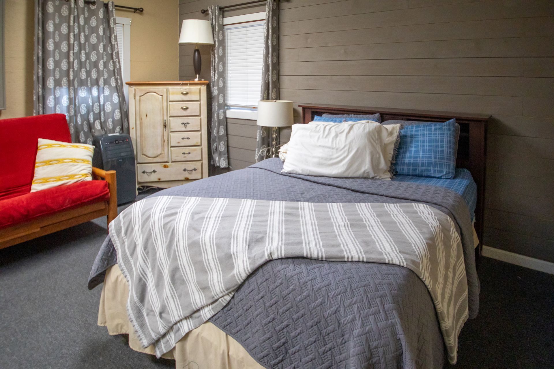Bedroom with a bed, dresser, futon, curtains, and lamps; neutral color palette.