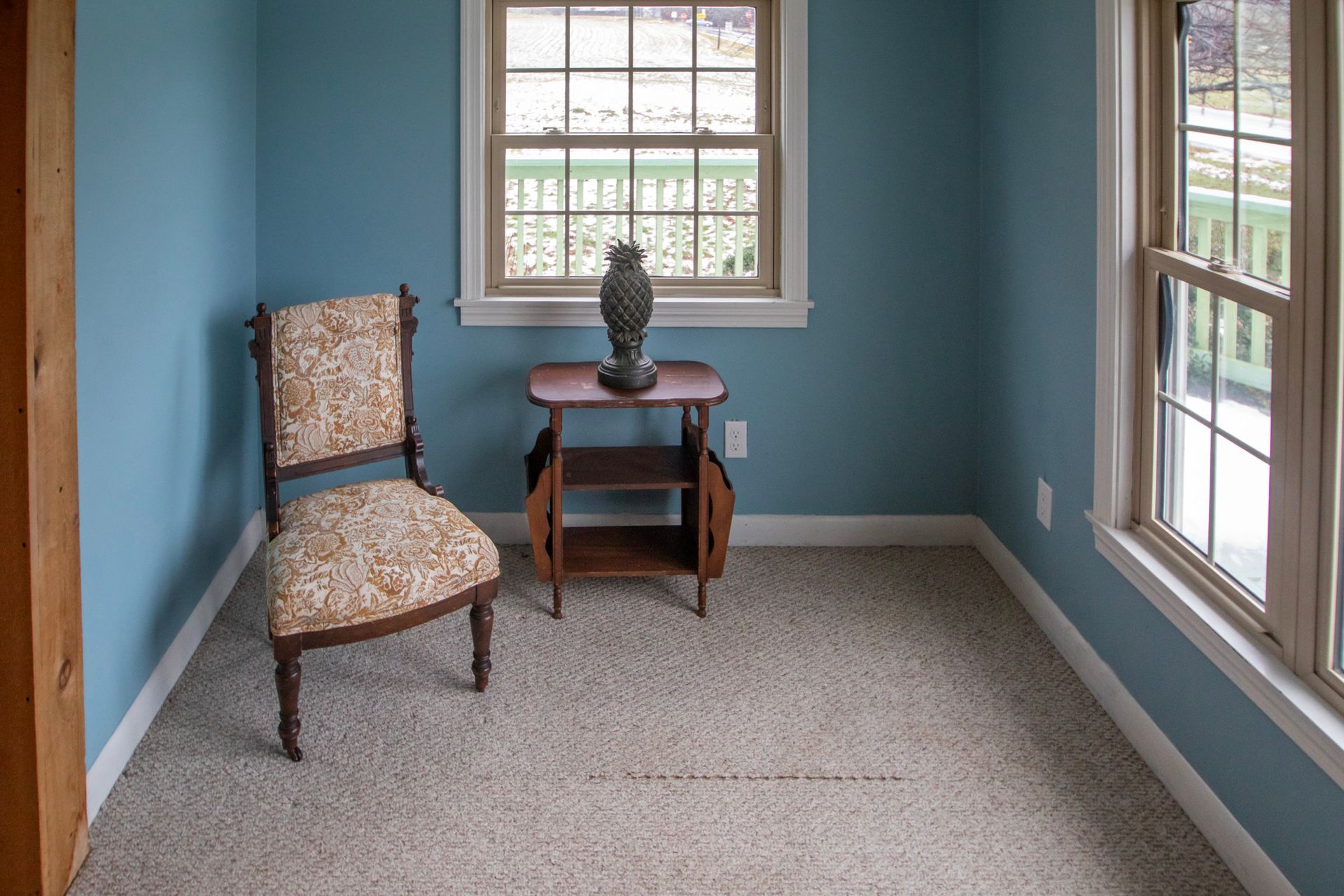 Small blue room with chair, small table, and windows. Beige carpet and white trim.