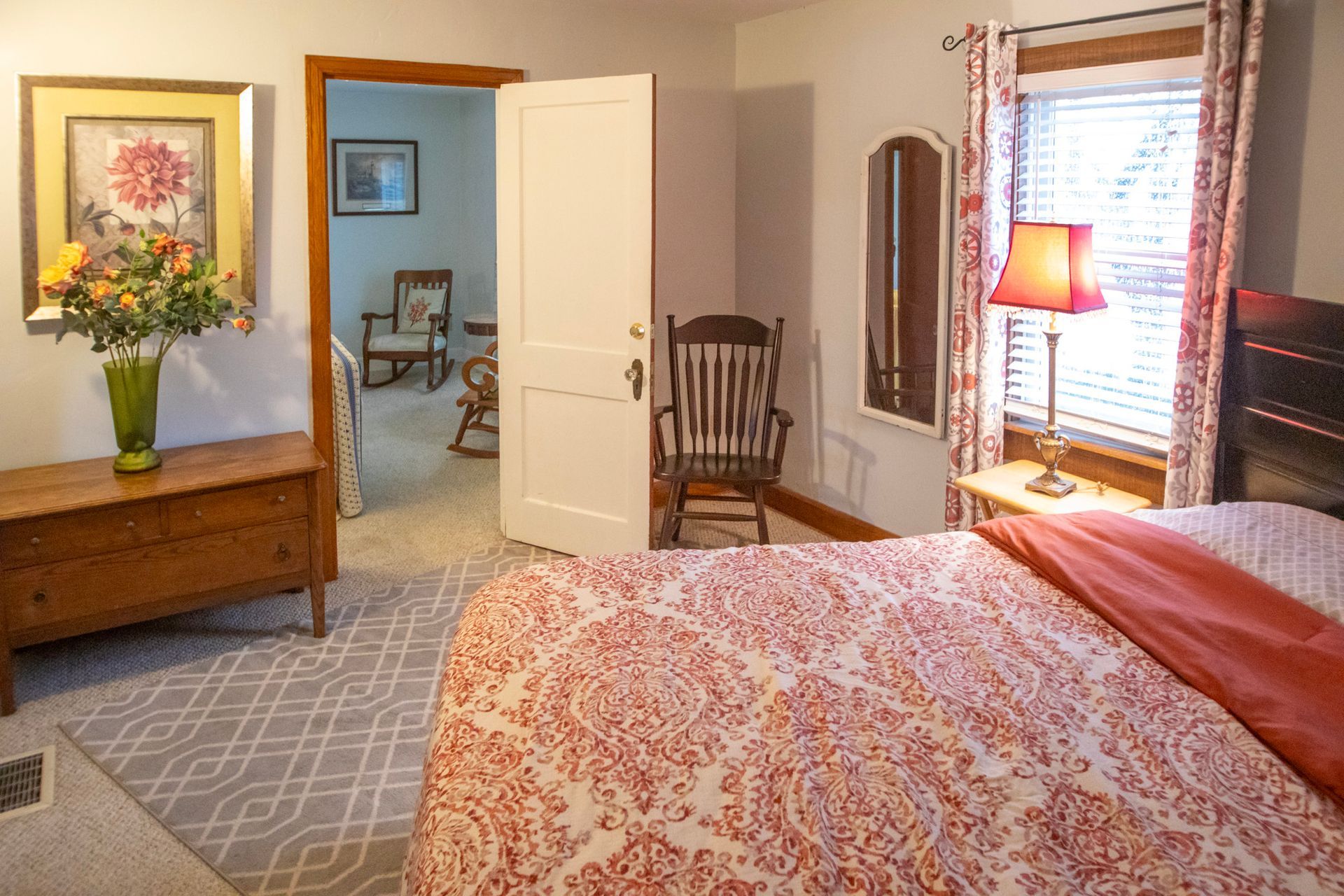 Bedroom with a bed, dresser, chair, mirror, and doorway leading to another room.