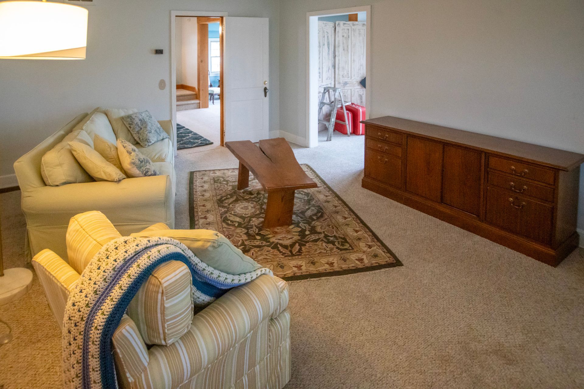 Living room with cream-colored sofas, wooden bench, and cabinet, with doors leading to other rooms.