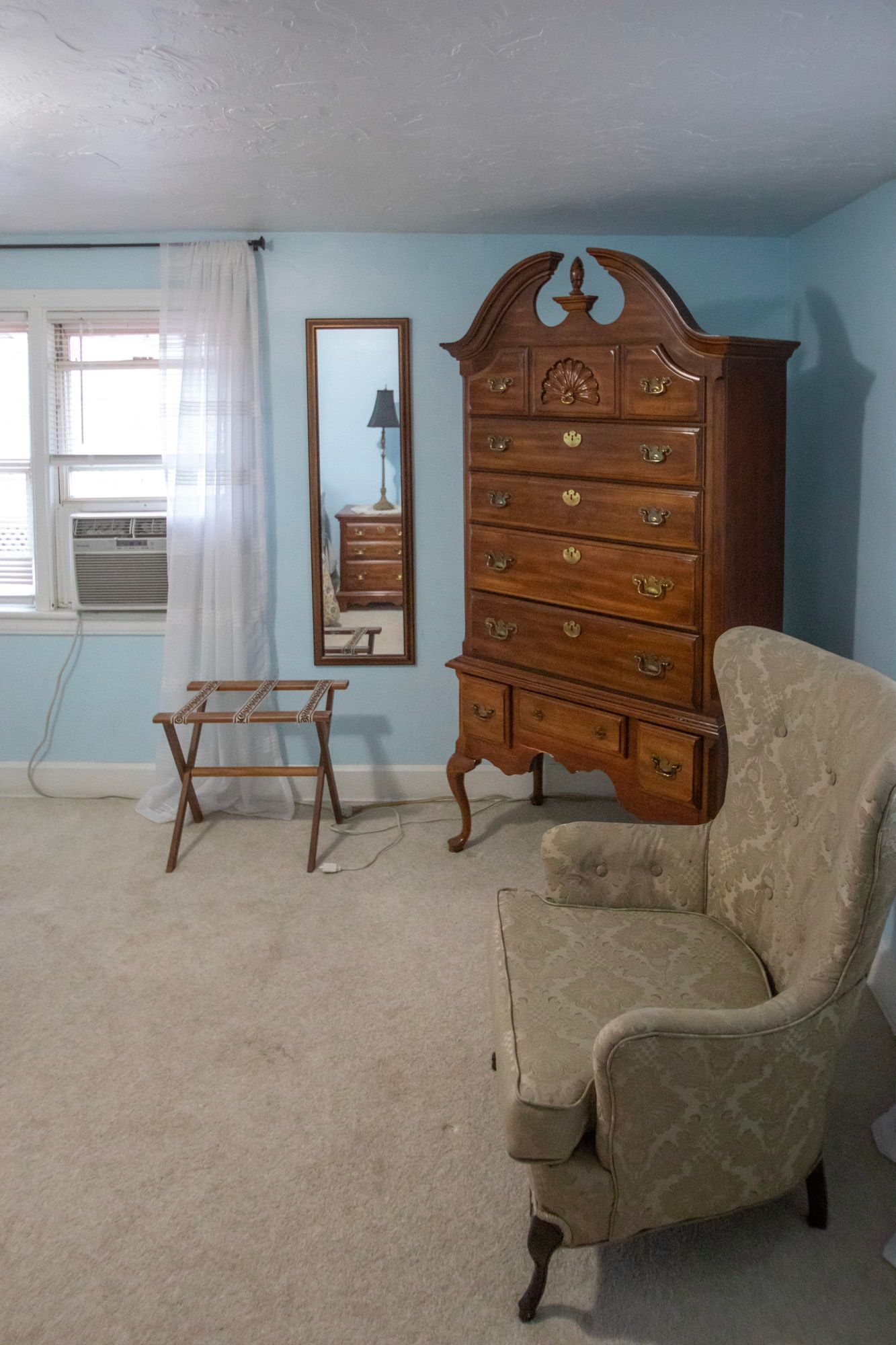 Bedroom with antique furniture: tall chest, wingback chair, mirror, and serving tray. Light blue walls, light carpet.