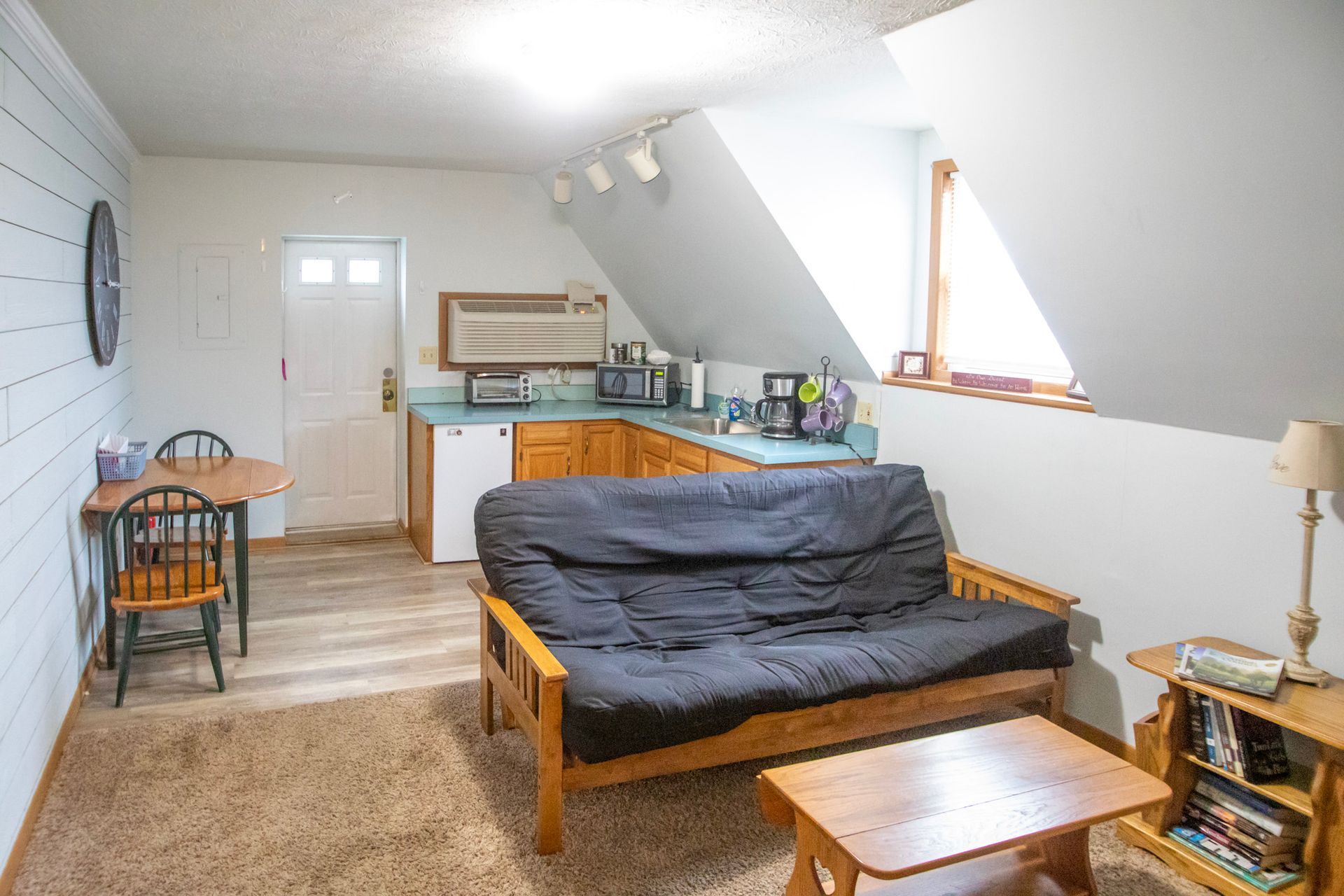 Small apartment with futon, kitchenette, and dining table. Neutral colors with sloped ceiling.
