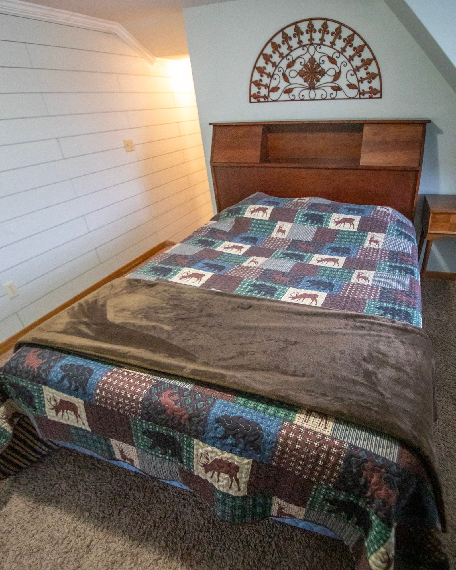 Bedroom with wood headboard, quilt, and brown blanket. White paneling on wall. Decorative metal art above headboard.