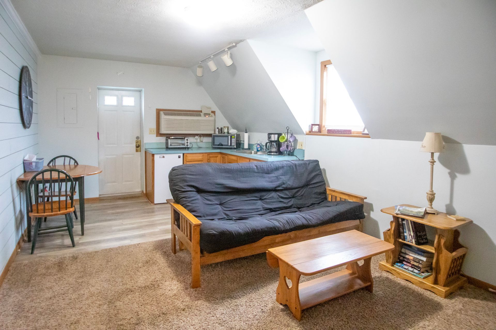 Small living space with futon, table, chairs, kitchenette, and slanted ceiling.