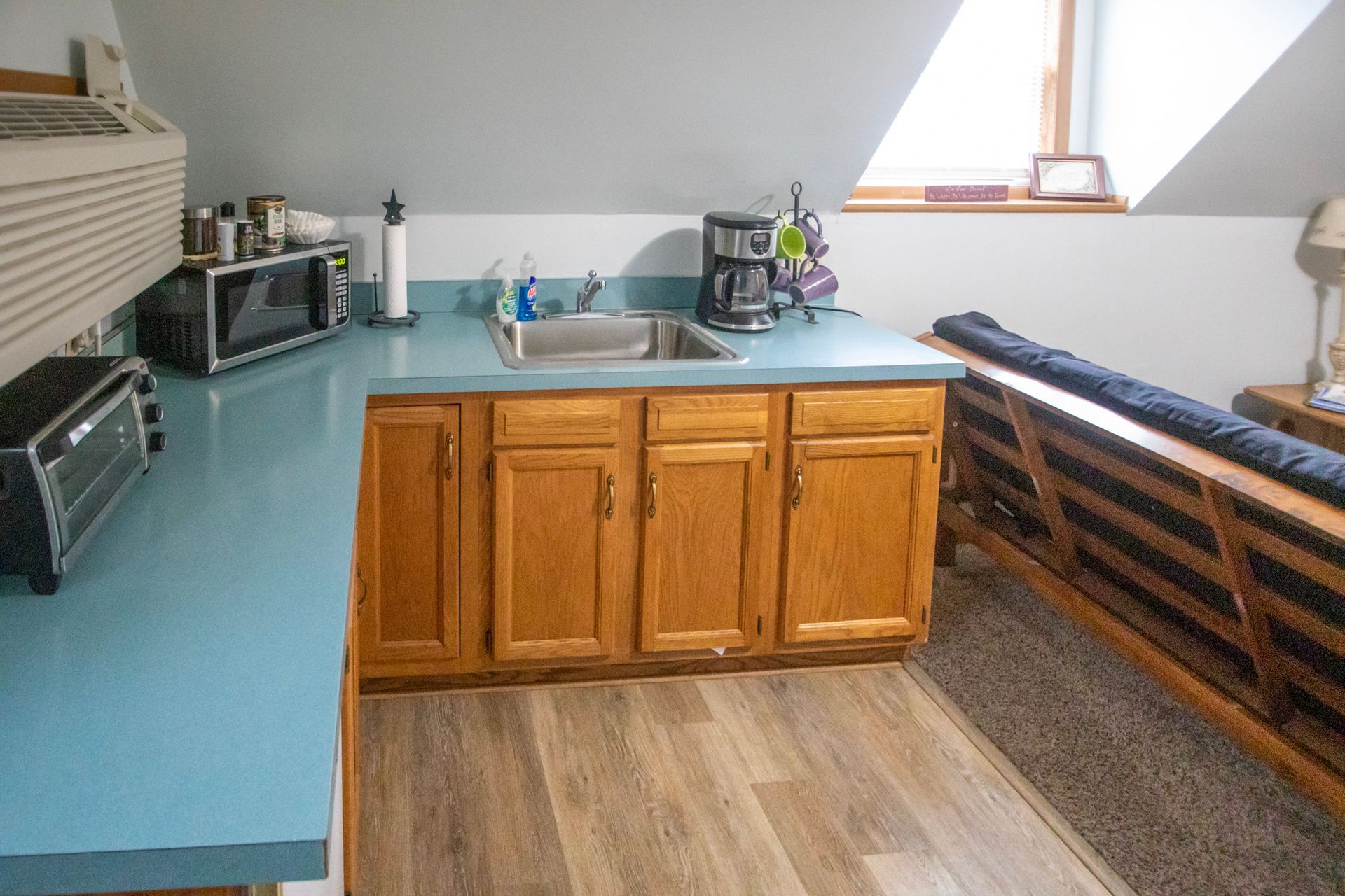 Small kitchenette with blue countertop, wooden cabinets, sink, microwave, toaster, and coffee maker.