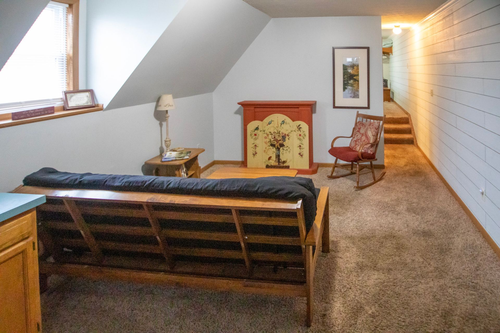 Cozy, carpeted room with angled ceiling, futon, fireplace, rocking chair, and stairs leading to another area.