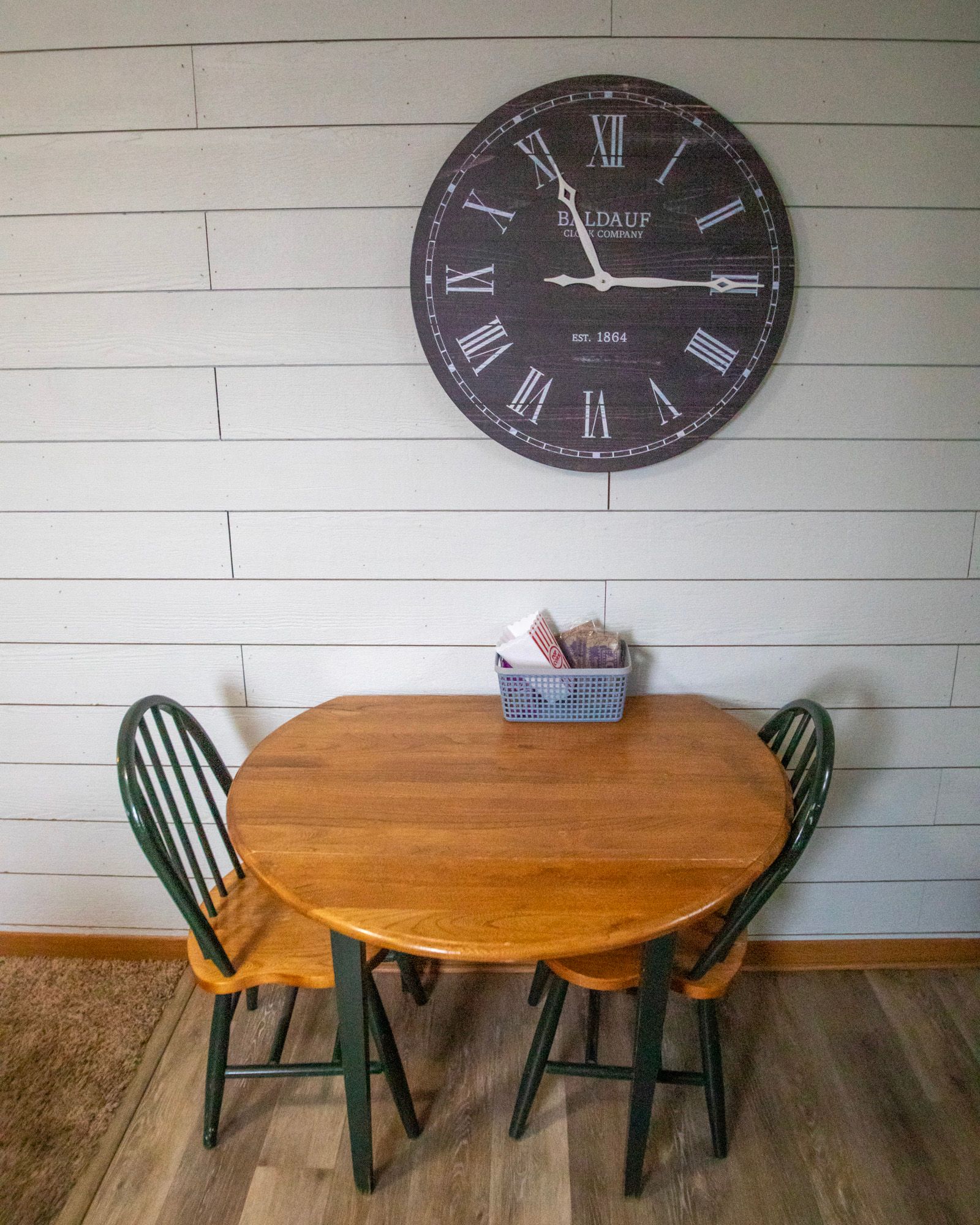 Wooden dining table with two chairs, under a large clock on a white wall.