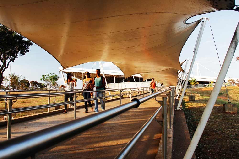 Pessoas caminhando sob um grande toldo com grade de metal ao longo de uma trilha.