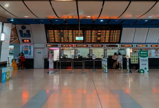 Bilheterias de estação de trem com um grande painel de partidas. Pessoas nos guichês sob um teto azul e branco.