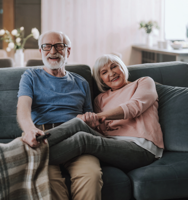 A couple sits on a grey sofa, holding hands while watching television with a remote in a comfortable home living room.