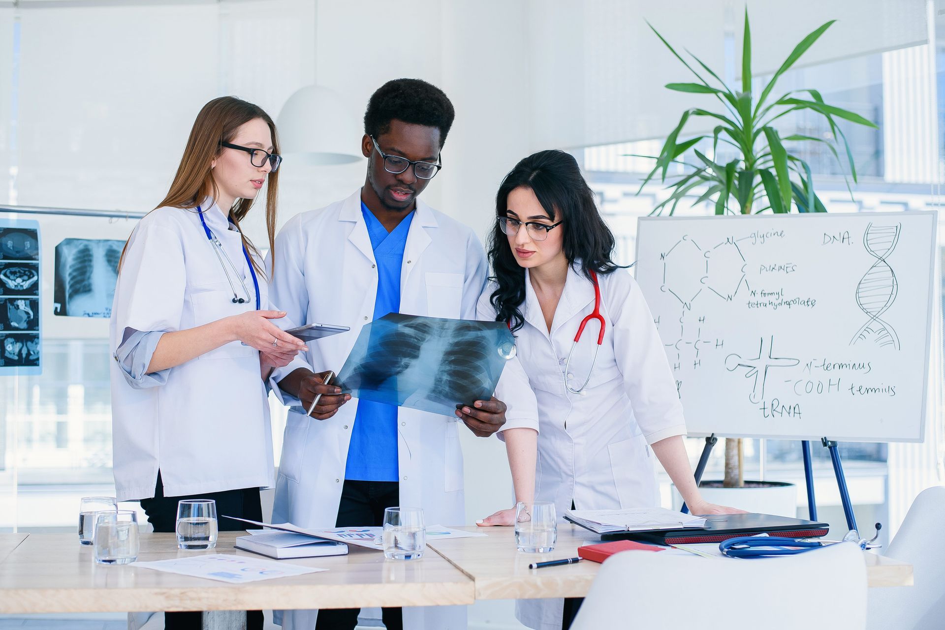 Three medical professionals in white coats analyze an X-ray in a clinical office setting with a whiteboard behind them.