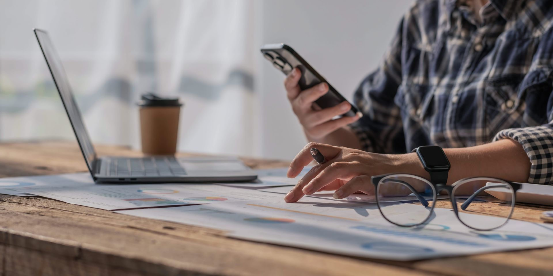 A person in a plaid shirt works at a wooden desk with a laptop, smartphone, pen, and glasses atop documents.