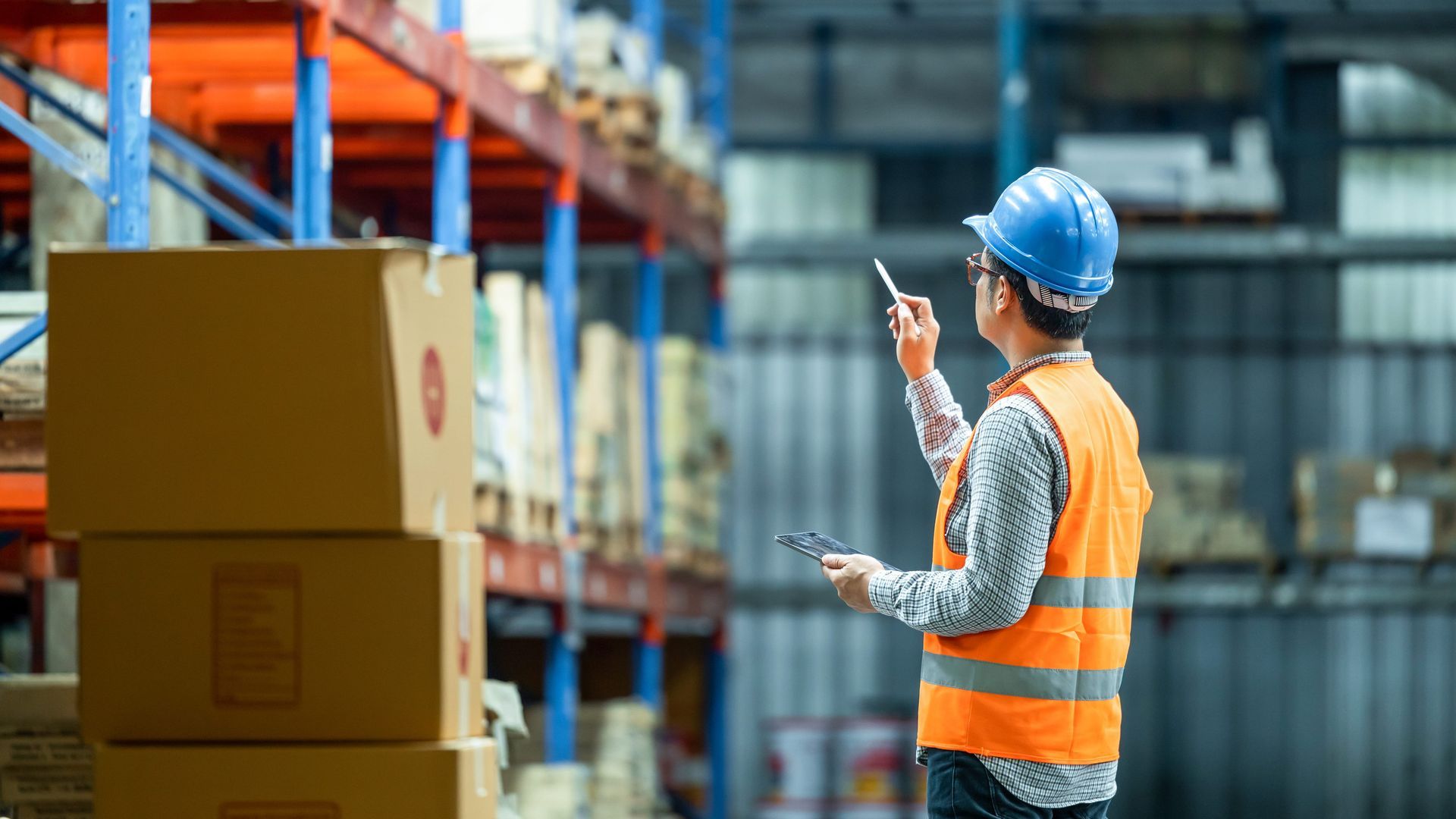 A warehouse worker in a hard hat and safety vest checks stock on a shelf while holding a pen and tablet.