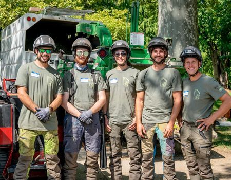 Five tree workers in green uniforms and helmets pose in front of equipment.