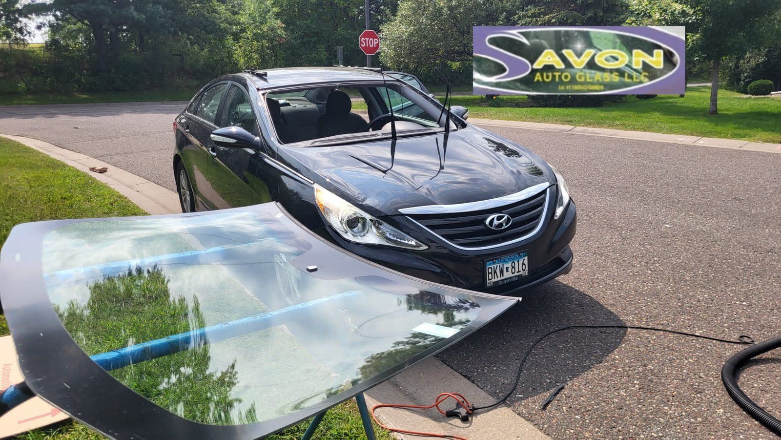 A black Hyundai Sonata with a new windshield being installed in a driveway by a company, Avon Auto Glass.