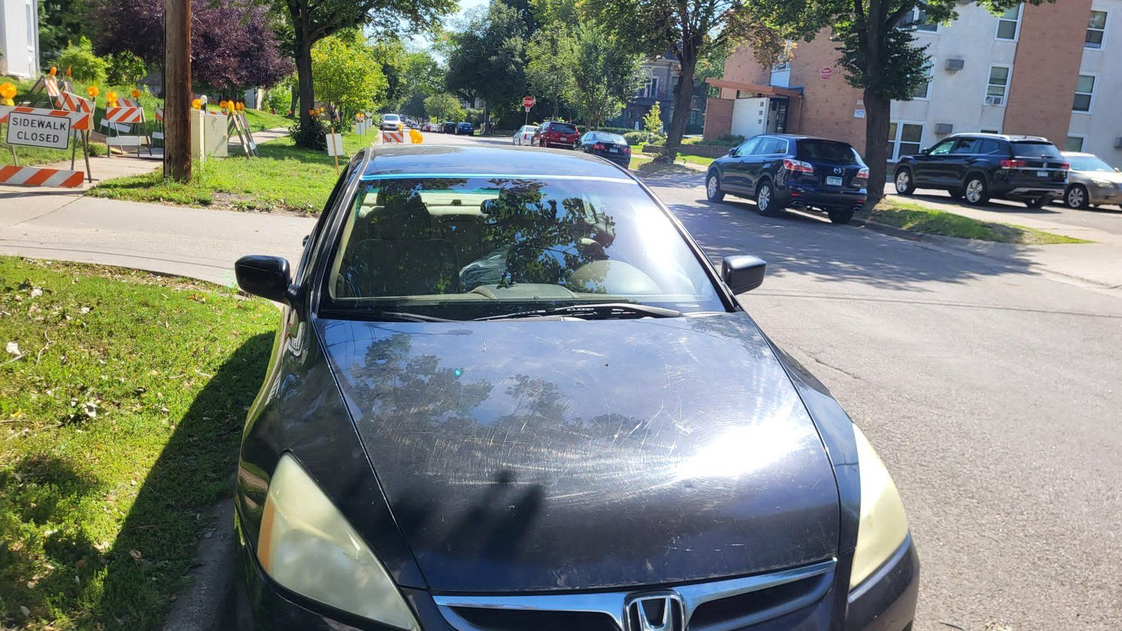 Black car parked on street; person visible in driver's seat. Trees and parked cars line the street in sunny weather.