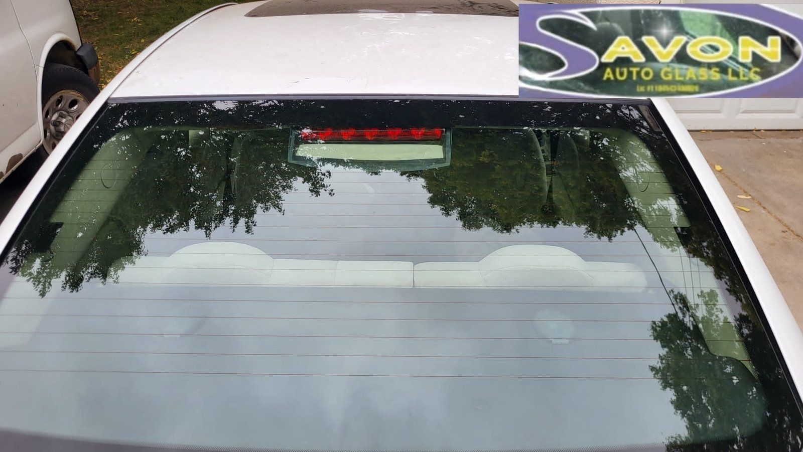White car with rear window showing adhesive residue, outdoors. Trees reflect in the glass.