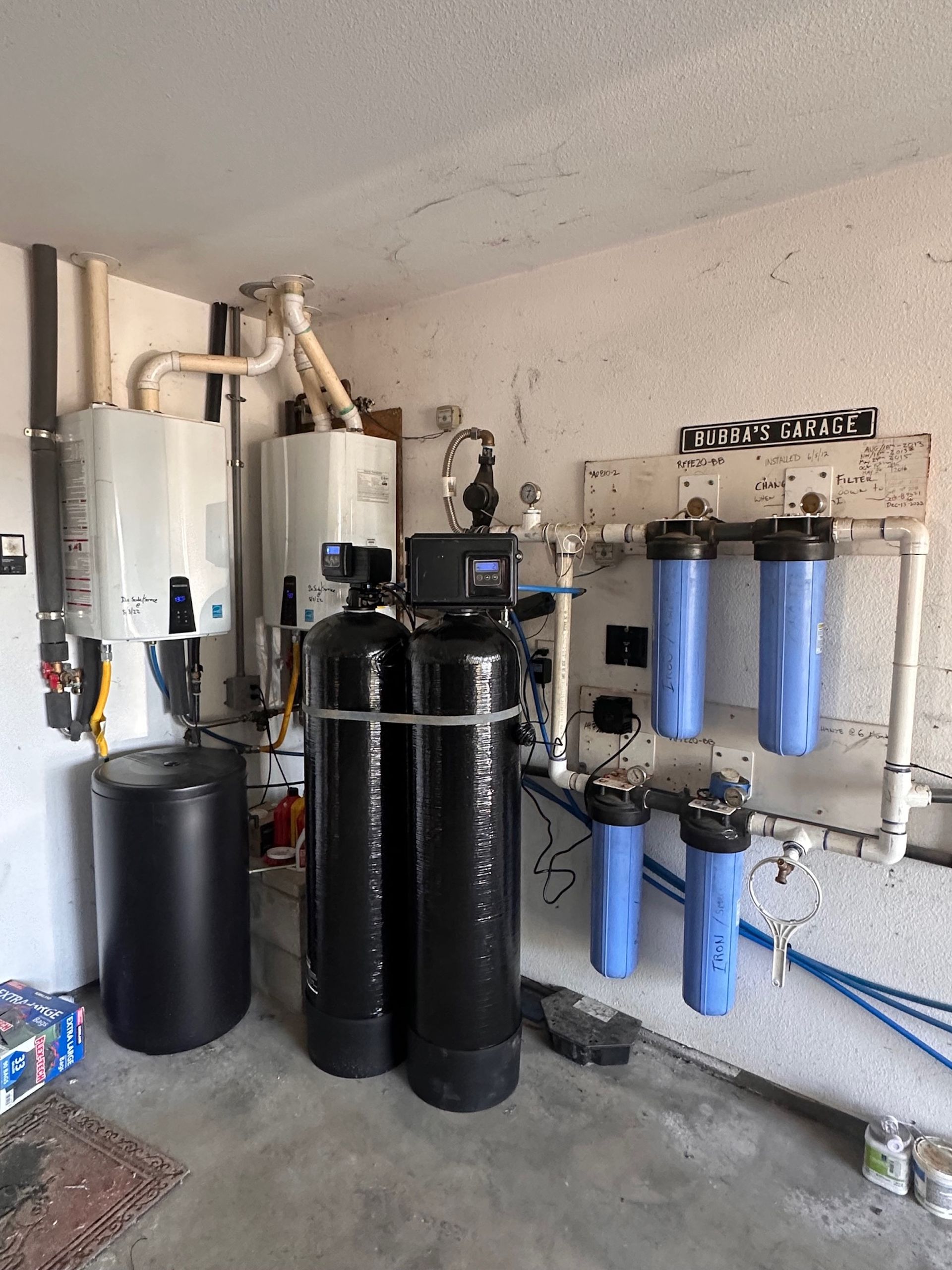 Water filtration and heating system in a utility room with black tanks, blue filters, and white pipes.