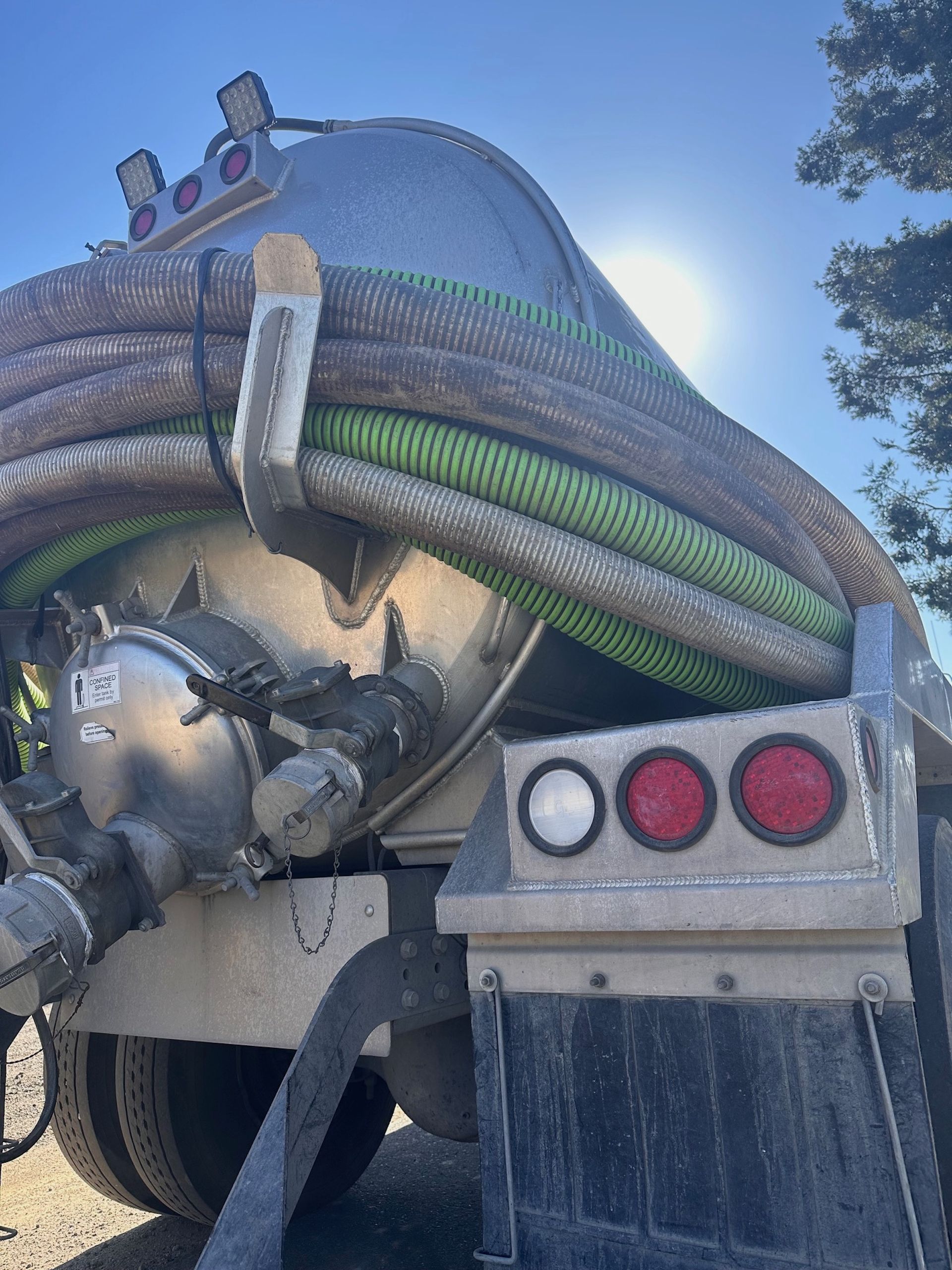 Rear view of a silver tanker truck with hoses.  Bright sun behind the truck, mud flaps, and red and white lights visible.
