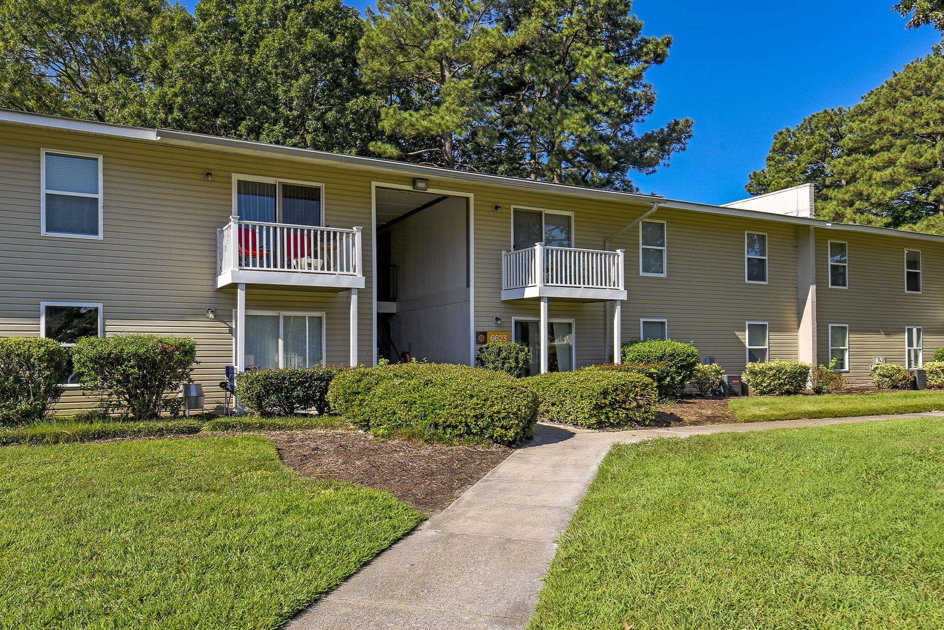 Exterior view of a beige apartment building with balconies, hedges, and a walkway at Chesapeake Pointe, offers  pet-friendly apartments in Chesapeake, VA