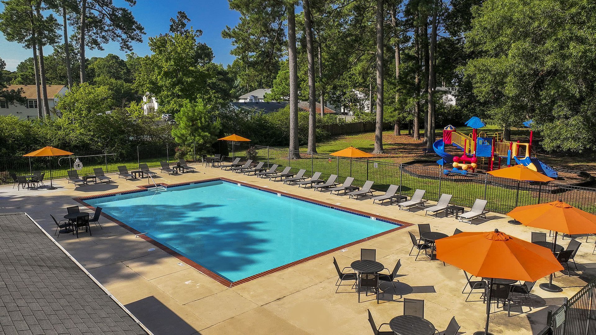Outdoor apartment pool area with lounge chairs and orange umbrellas; a playground visible beyond at Chesapeake Pointe, offers  pet-friendly apartments in Chesapeake, VA