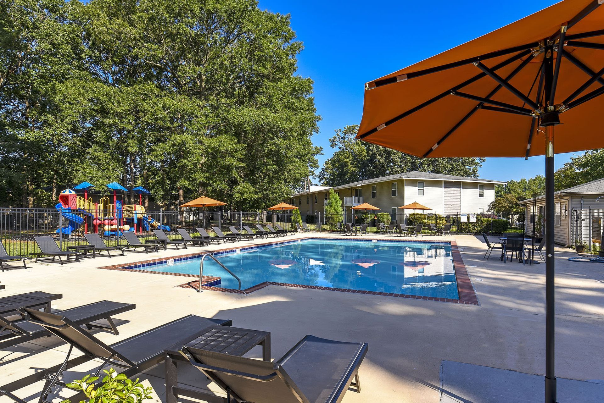 Outdoor apartment community pool with lounge chairs, umbrellas, and a playground in the background at Chesapeake Pointe, offers  pet-friendly apartments in Chesapeake, VA