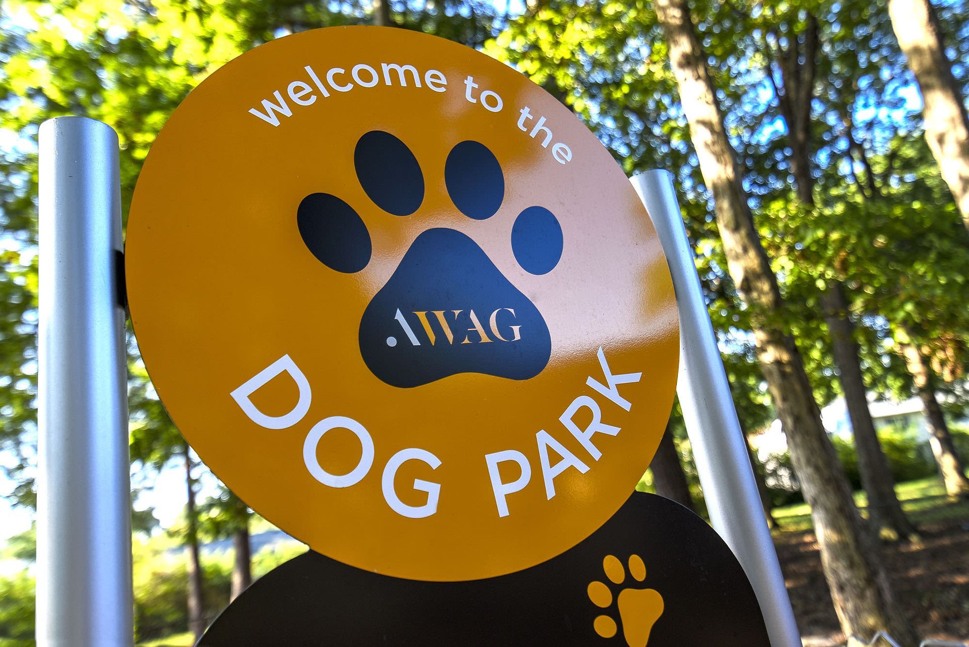 Bright yellow round dog park sign with a large black paw print and white text, outdoors among trees at Chesapeake Pointe, offers  pet-friendly apartments in Chesapeake, VA