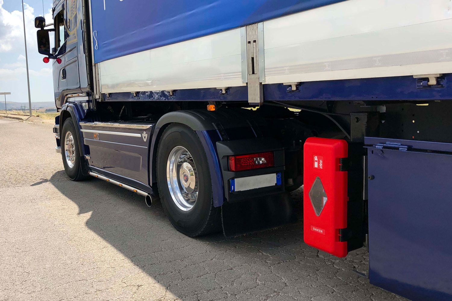 Semi-camion bleu avec un conteneur rouge sur le côté, stationné à l'extérieur au soleil.