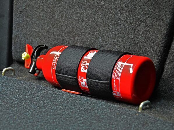 Extincteur rouge fixé dans un véhicule, maintenu par des sangles noires, reposant sur un tapis gris.