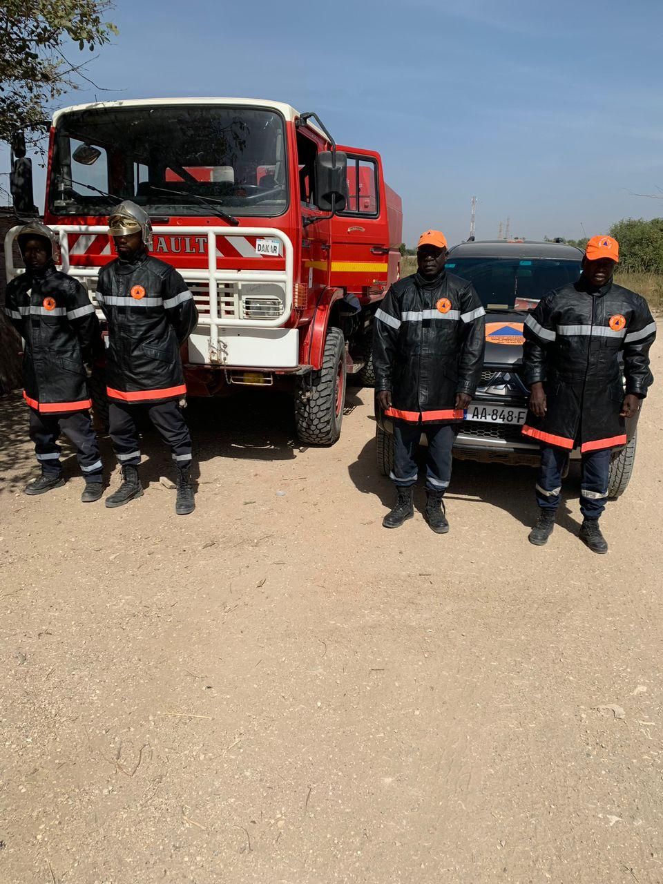 Les pompiers en uniforme se tiennent près d'un camion de pompiers rouge et d'un véhicule noir, journée ensoleillée.