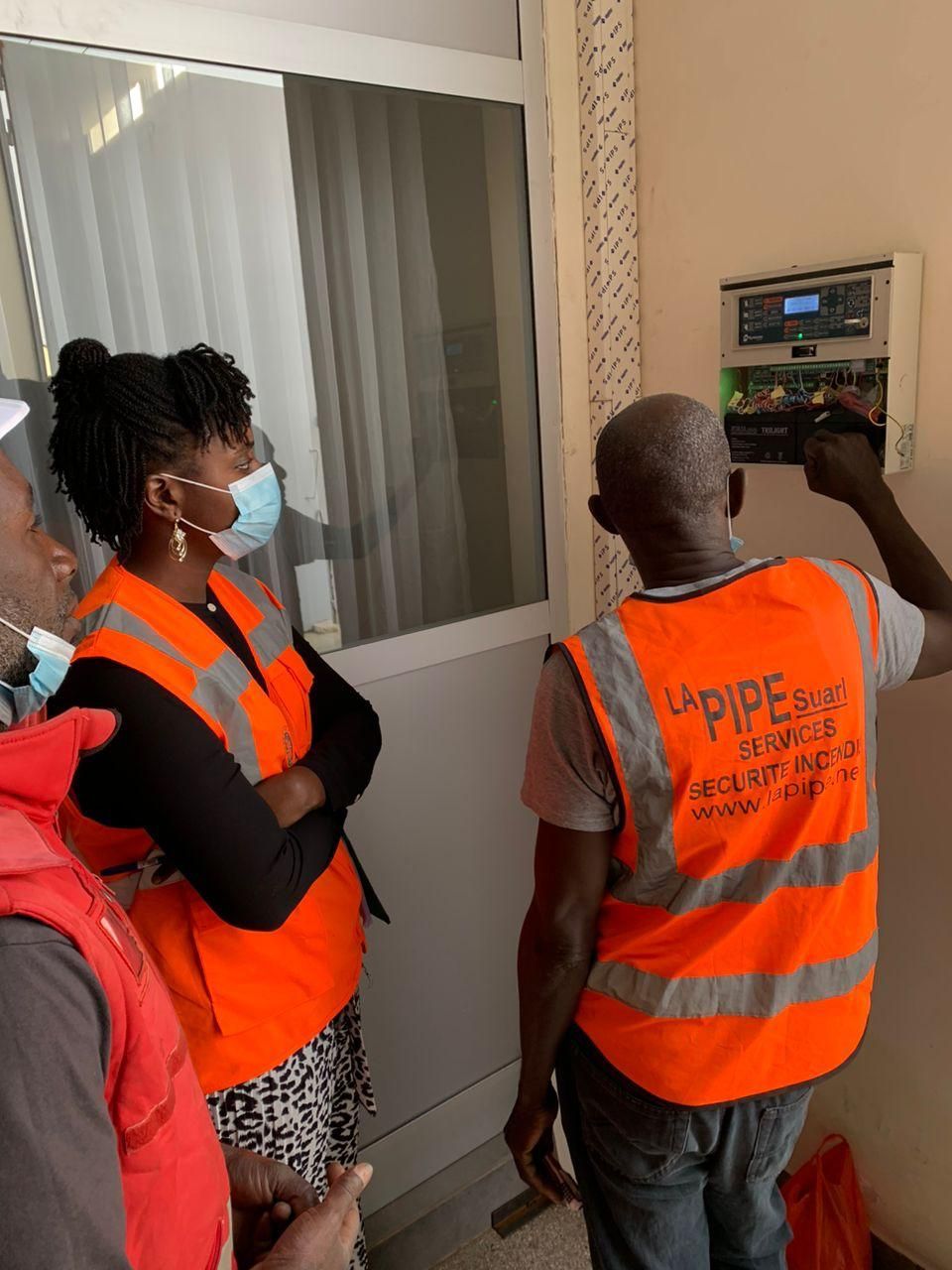 Trois personnes en gilet orange examinent un panneau électrique sur un mur blanc. L'une d'elles pointe du doigt.