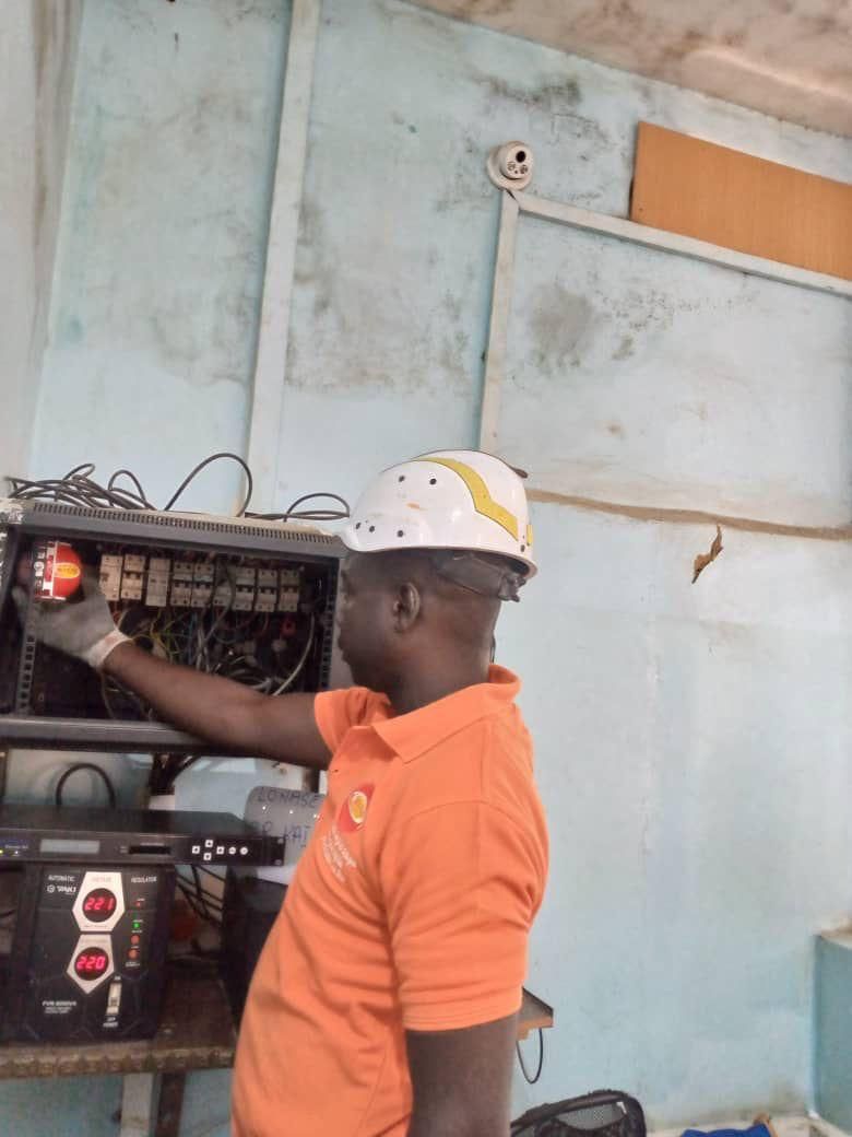 Un homme en chemise orange et casque travaille sur un panneau électrique à l'intérieur.