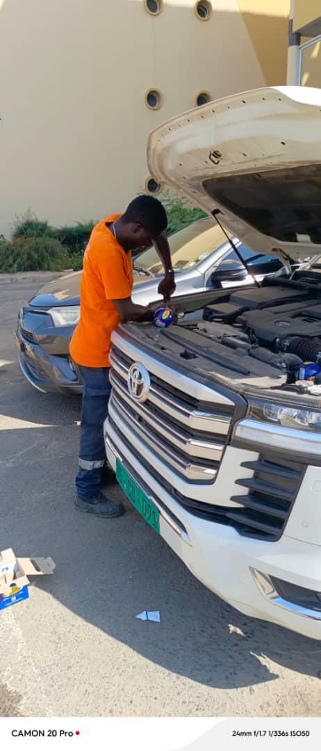 Un mécanicien en chemise orange travaille sur une voiture blanche, capot ouvert. Une voiture grise est garée à côté.