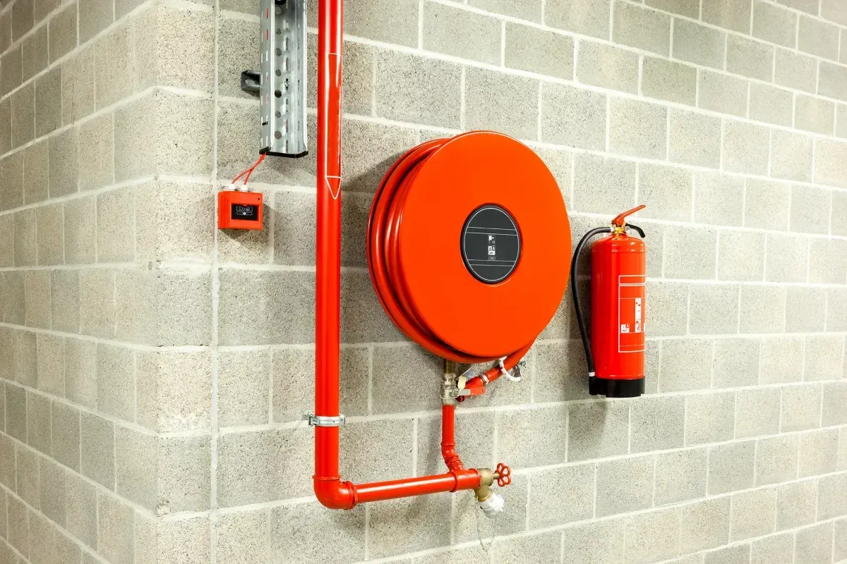 Enrouleur de tuyau d'incendie et extincteur sur un mur en blocs de béton, avec tuyauterie rouge et un petit boîtier d'alarme.
